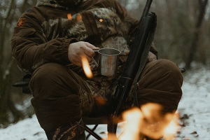 man sitting at campfire with coffee and gun