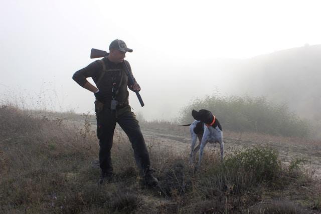 man holding gun over shoulder with bird dog in field